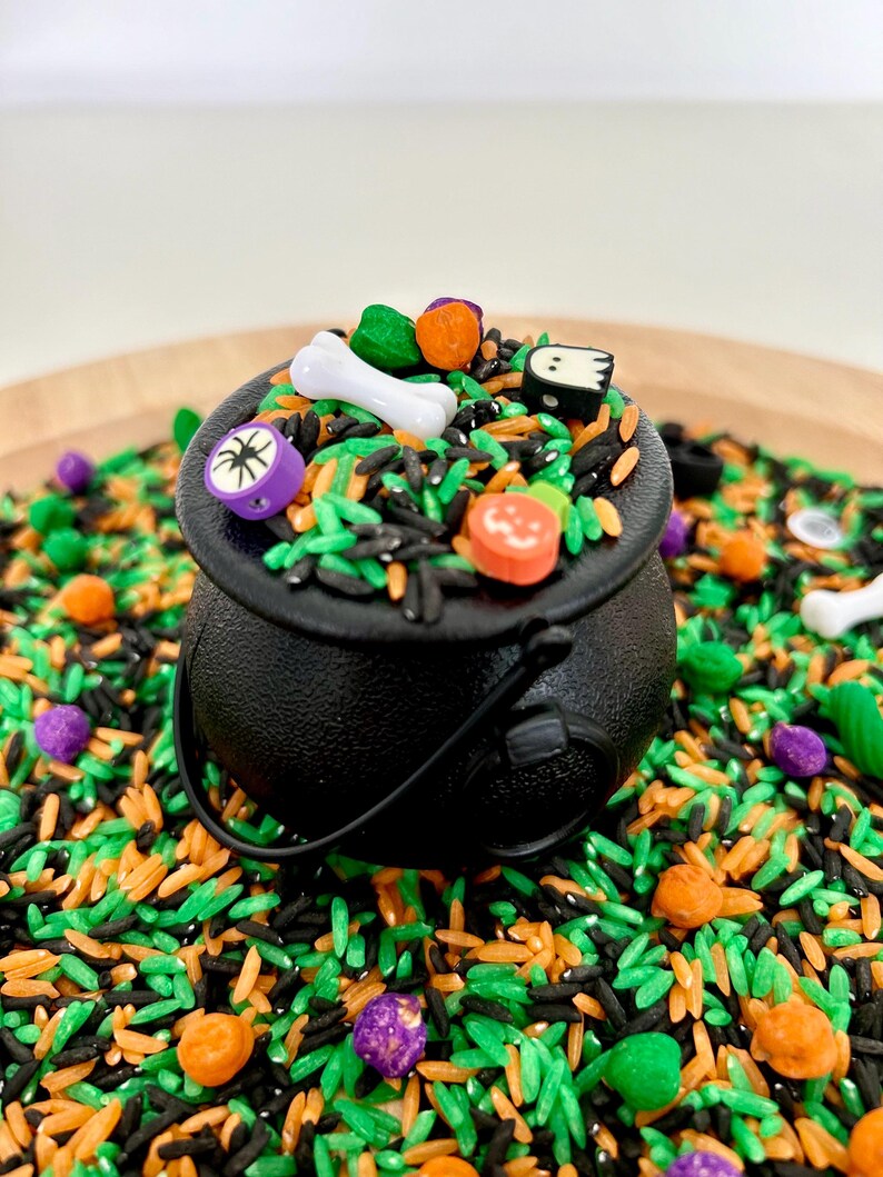 May include: A black cauldron filled with colorful Halloween-themed sensory rice. The cauldron is surrounded by green, orange, black, and purple rice, with small decorative pieces like bones and pumpkins. The cauldron has a black handle.