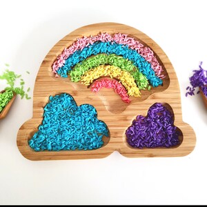 May include: A wooden rainbow-shaped tray filled with coloured rice in pink, blue, green, yellow, and red. Two cloud-shaped compartments hold blue and purple rice. Wooden scoops with green and purple rice are on either side.