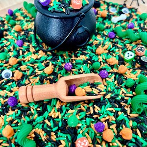 May include: A Halloween-themed sensory bin with a black cauldron filled with colorful rice, small pumpkins, ghosts, and other festive decorations. A wooden scoop is filled with purple and orange beads. Green, orange, and black rice create a vibrant background.
