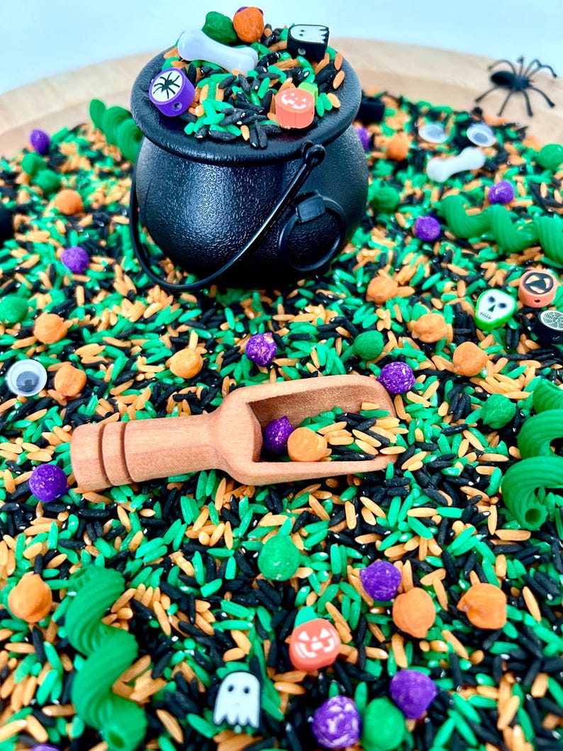May include: A Halloween-themed sensory bin with a black cauldron filled with colorful rice, small pumpkins, ghosts, and other festive decorations. A wooden scoop is filled with purple and orange beads. Green, orange, and black rice create a vibrant background.