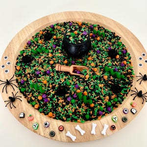 May include: A wooden tray filled with a sensory bin for Halloween. The bin contains green, black, orange, and purple colored rice, a black cauldron, a wooden scoop, plastic spiders, and Halloween-themed beads.