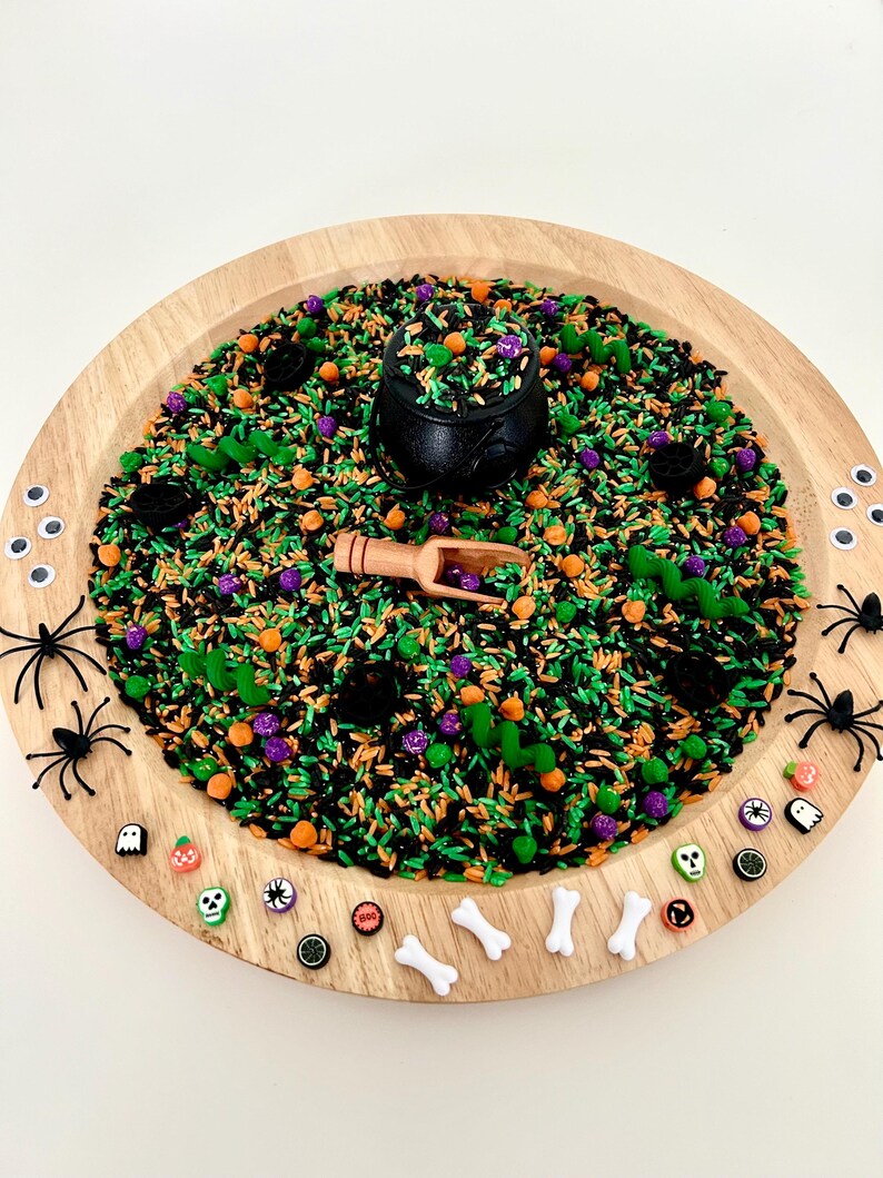 May include: A wooden tray filled with a sensory bin for Halloween. The bin contains green, black, orange, and purple colored rice, a black cauldron, a wooden scoop, plastic spiders, and Halloween-themed beads.