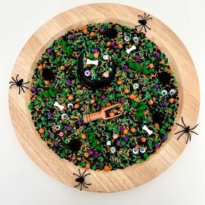 May include: A wooden tray filled with a sensory bin for Halloween. The bin contains black, green, orange, and purple sprinkles, along with small Halloween-themed decorations like spiders, bones, and eyeballs. A small wooden scoop is included.