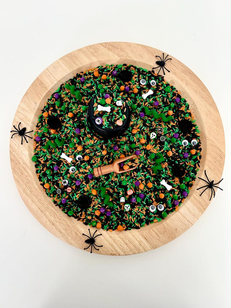 May include: A wooden tray filled with a sensory bin for Halloween. The bin contains black, green, orange, and purple sprinkles, along with small Halloween-themed decorations like spiders, bones, and eyeballs. A small wooden scoop is included.