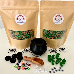 May include: Two brown paper bags with clear windows, each with a rainbow sticker that says "Messy Munchkins". A black cauldron, spiders, and various Halloween-themed embellishments are arranged on a white surface. Includes a wooden scoop, bones, and googly eyes.