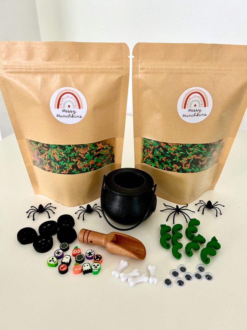 May include: Two brown paper bags with clear windows, each with a rainbow sticker that says "Messy Munchkins". A black cauldron, spiders, and various Halloween-themed embellishments are arranged on a white surface. Includes a wooden scoop, bones, and googly eyes.