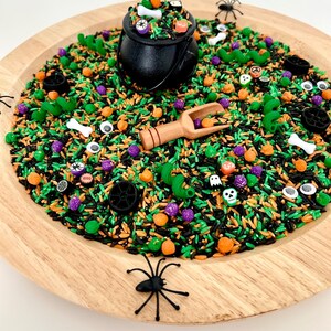 May include: A Halloween-themed sensory bin with black, orange, green, and purple rice. A black cauldron is filled with decorative items, including bones and pumpkins. A wooden scoop and plastic spiders are also included. The bin is on a round wooden tray.