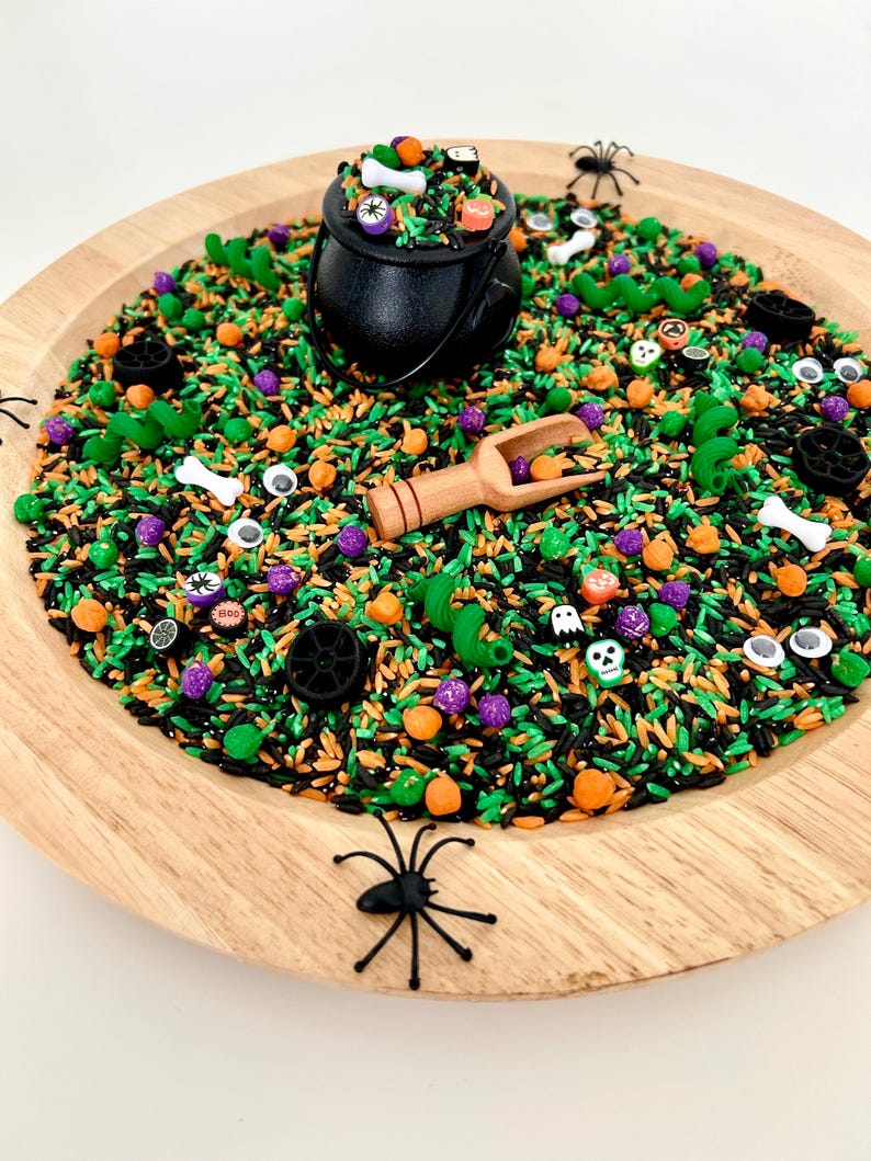 May include: A Halloween-themed sensory bin with black, orange, green, and purple rice. A black cauldron is filled with decorative items, including bones and pumpkins. A wooden scoop and plastic spiders are also included. The bin is on a round wooden tray.