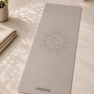 Op de afbeelding: Een rechthoekige lichtgrijze yogamat met een centraal lijntekening-ontwerp van een maansikkel omringd door stralende lijnen en kleine sterren. Het woord "mayuna" staat in een donkergrijs schreefloos lettertype bij de onderrand.