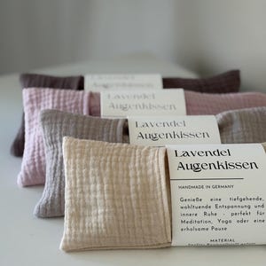Könnte beinhalten: Ein Stapel aus vier rechteckigen Augenkissen aus strukturiertem Stoff in den Farben Beige, Grau, Rosa und Dunkelbraun. Jedes Kissen hat ein weißes Papieretikett mit dem Text Lavendel Augenkissen und weiterem deutschen Text.
