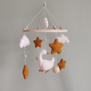 May include: A baby mobile featuring a white goose, mustard-colored stars, white clouds, and wooden beads. The mobile is suspended from a wooden ring and is designed for a nursery or children's room.
