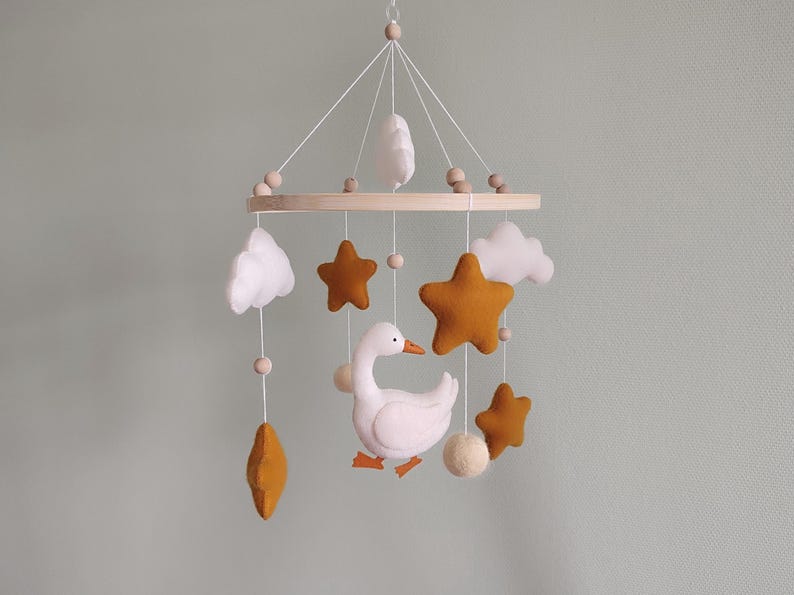May include: A baby mobile featuring a white goose, mustard-colored stars, white clouds, and wooden beads. The mobile is suspended from a wooden ring and is designed for a nursery or children's room.