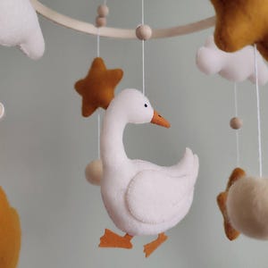May include: A handmade baby mobile featuring a white goose, mustard yellow stars, white clouds, and wooden beads. The mobile is suspended from a wooden hoop with delicate strings, creating a whimsical nursery decoration.