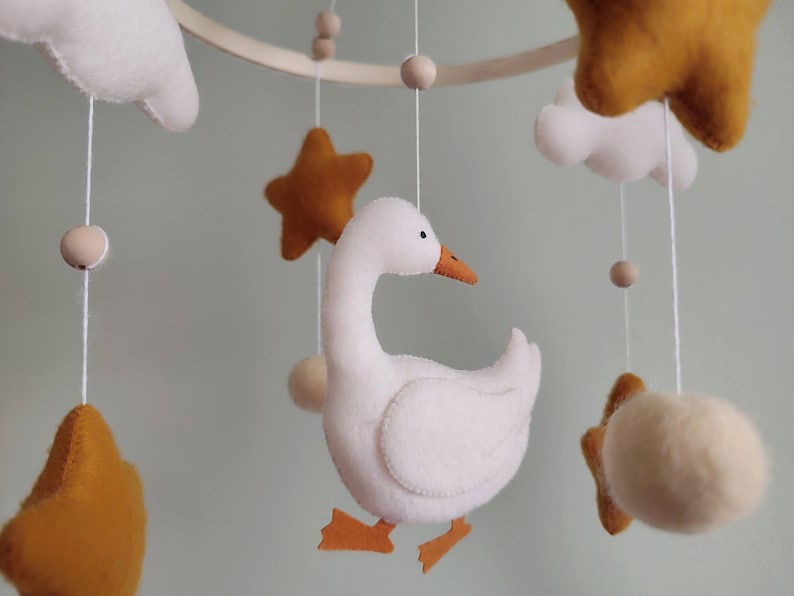 May include: A handmade baby mobile featuring a white goose, mustard yellow stars, white clouds, and wooden beads. The mobile is suspended from a wooden hoop with delicate strings, creating a whimsical nursery decoration.