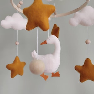 May include: A handmade baby mobile featuring a white goose with orange beak and feet, surrounded by mustard-colored stars and white clouds. The mobile is suspended from a wooden hoop with wooden beads, creating a whimsical nursery decoration.
