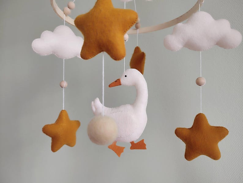 May include: A handmade baby mobile featuring a white goose with orange beak and feet, surrounded by mustard-colored stars and white clouds. The mobile is suspended from a wooden hoop with wooden beads, creating a whimsical nursery decoration.
