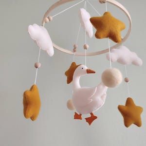 May include: A handmade baby mobile featuring a wooden hoop with hanging felt stars, clouds, and a goose. The stars are mustard yellow, the clouds are white, and the goose is white with orange feet and a beak. The mobile is designed for a nursery.