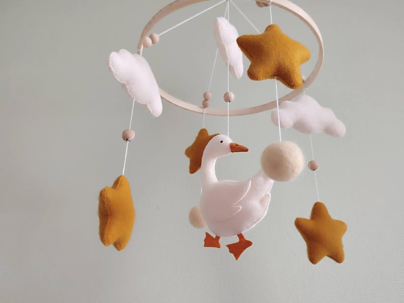 May include: A handmade baby mobile featuring a wooden hoop with hanging felt stars, clouds, and a goose. The stars are mustard yellow, the clouds are white, and the goose is white with orange feet and a beak. The mobile is designed for a nursery.