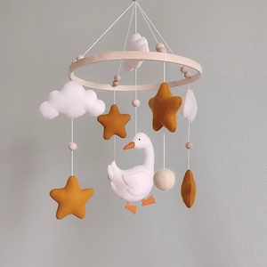 May include: A baby mobile featuring a wooden ring with hanging felt shapes. Includes white clouds, mustard-colored stars, and a white goose with orange feet and beak. The mobile is suspended by white string and wooden beads.