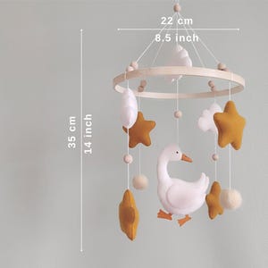May include: A baby mobile with a wooden ring, featuring a white goose, mustard-colored stars, white clouds, and felt balls. The mobile is 8.5 inches wide and 14 inches long, designed for a nursery.
