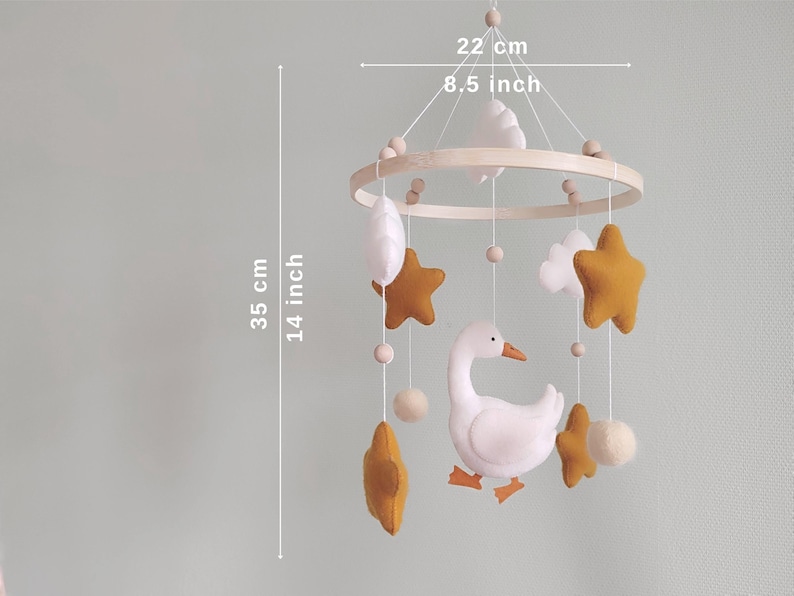 May include: A baby mobile with a wooden ring, featuring a white goose, mustard-colored stars, white clouds, and felt balls. The mobile is 8.5 inches wide and 14 inches long, designed for a nursery.