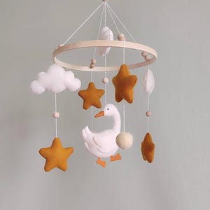 May include: A baby mobile featuring a wooden ring with hanging felt shapes. Includes mustard yellow stars, white clouds, a white goose with orange feet, and a white ball. The mobile is suspended by white strings.