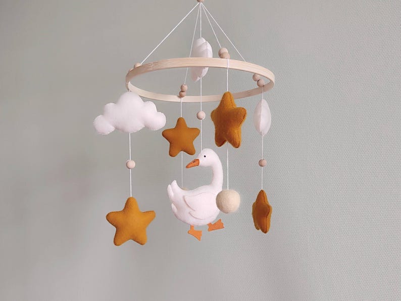 May include: A baby mobile featuring a wooden ring with hanging felt shapes. Includes mustard yellow stars, white clouds, a white goose with orange feet, and a white ball. The mobile is suspended by white strings.