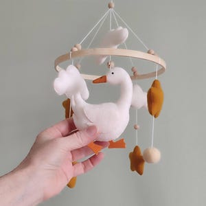 May include: A handmade baby mobile featuring a white goose with orange feet and beak, surrounded by white clouds, mustard-colored stars, and a wooden ring. The mobile is suspended by white strings and wooden beads.