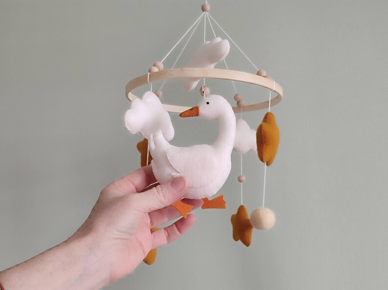 May include: A handmade baby mobile featuring a white goose with orange feet and beak, surrounded by white clouds, mustard-colored stars, and a wooden ring. The mobile is suspended by white strings and wooden beads.