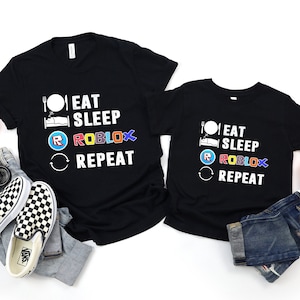 May include: Two black t-shirts with the text "Eat Sleep Roblox Repeat" in white. The word "Roblox" is in rainbow colors. The shirts are on a white background with some Halloween decorations.