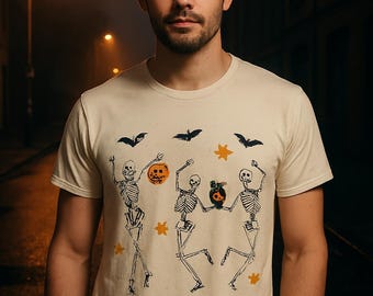 May include: A cream-coloured t-shirt with a Halloween-themed design. The graphic features dancing skeletons, bats, and stars in orange and black. The shirt is made of a soft material, suitable for casual wear.