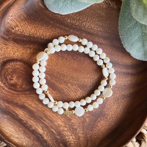 May include: A double-strand bracelet featuring creamy white beads and gold accents. The bracelet includes a small gold cross charm and is displayed on a wooden tray. The beads are round and oval-shaped.