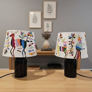 Op de afbeelding: Twee decoratieve tafellampen met zwarte voetstukken en witte kappen. De kappen hebben kleurrijke geborduurde ontwerpen van herten, vogels en bloemmotieven. De lampen staan op een houten tafel.