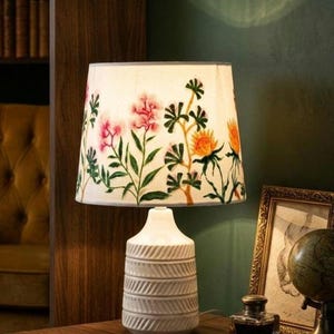 Könnte beinhalten: Eine weiße Tischlampe mit einem floral gemusterten Lampenschirm mit rosa und orangefarbenen Blumen. Die Lampe hat einen strukturierten, cremefarbenen Sockel und steht auf einer dunklen Holzoberfläche. Ein gerahmtes Bild und ein Globus sind im Hintergrund zu sehen.