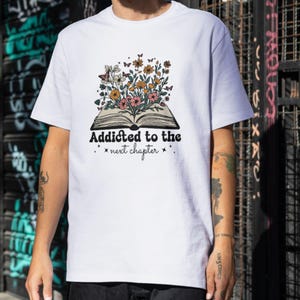 以下が含まれることがあります： 開いた本、花、蝶のデザインが施された白いTシャツ。「Addicted to the next chapter」の文字が本の下にプリントされています。デザインにはピンク、黄色、オレンジの花が含まれています。