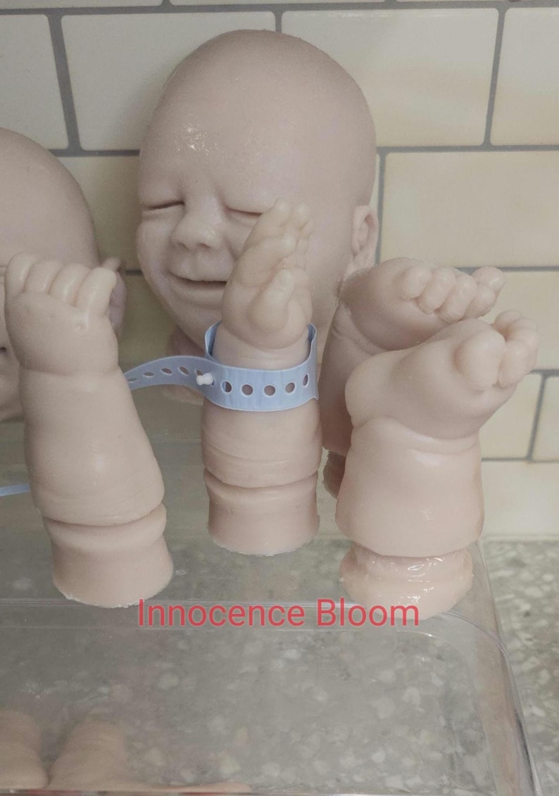 Pu&ograve; includere: Un kit per bambola reborn in vinile dalla pelle chiara composto da una testa con occhi chiusi, due braccia e due gambe. Un braccio indossa un braccialetto identificativo ospedaliero in plastica azzurra. Il testo Innocence Bloom &egrave; in rosso.