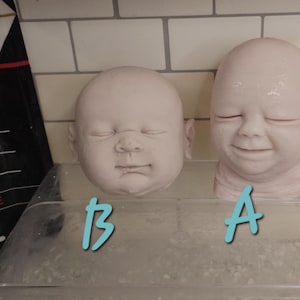 Pu&ograve; includere: Due teste di bambola in vinile non dipinte, etichettate A e B in testo azzurro. La testa A presenta un'espressione sorridente con gli occhi chiusi, mentre la testa B mostra un'espressione neutra con gli occhi chiusi. Entrambe le teste sono lisce e pallide.