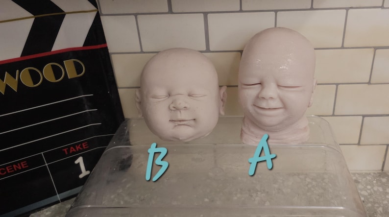 Pu&ograve; includere: Due teste di bambola in vinile non dipinte, etichettate A e B in testo azzurro. La testa A presenta un'espressione sorridente con gli occhi chiusi, mentre la testa B mostra un'espressione neutra con gli occhi chiusi. Entrambe le teste sono lisce e pallide.