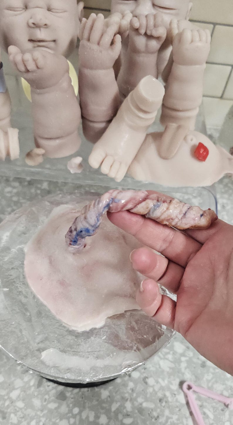 Pu&ograve; includere: Un modello in silicone di cordone ombelicale e placenta per bambole, caratterizzato da un cordone ritorto testurizzato rosa pallido con dettagli di vene blu, attaccato a una base placentare piatta, rotonda e color carne, vicino ad arti di bambola.