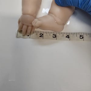 Pu&ograve; includere: Una coppia di arti per bambole in vinile di colore chiaro, composti da un braccio e un piede, posizionati contro un metro a nastro bianco che indica una lunghezza di circa 12,7 cm per l'insieme.