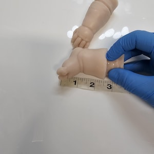 Pu&ograve; includere: Una coppia di arti per bambola in vinile di colore chiaro, composti da un braccio e un piede. Un metro a nastro bianco &egrave; posizionato contro il piede, indicando una lunghezza di circa 7,6 cm dal tallone alla zona delle dita del pezzo.