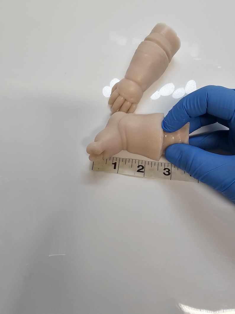 Pu&ograve; includere: Una coppia di arti per bambola in vinile di colore chiaro, composti da un braccio e un piede. Un metro a nastro bianco &egrave; posizionato contro il piede, indicando una lunghezza di circa 7,6 cm dal tallone alla zona delle dita del pezzo.