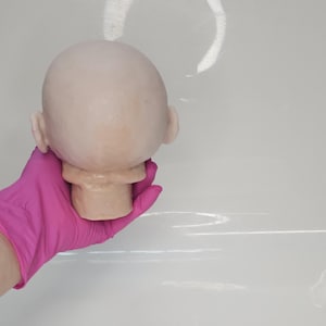 Pu&ograve; includere: Una testa di bambola calva e pallida con un cranio liscio e arrotondato e orecchie visibili. La testa &egrave; scolpita con un collo e una mascella definiti, caratterizzata da un tono di pelle chiaro con sottili variazioni di colore sul vinile.