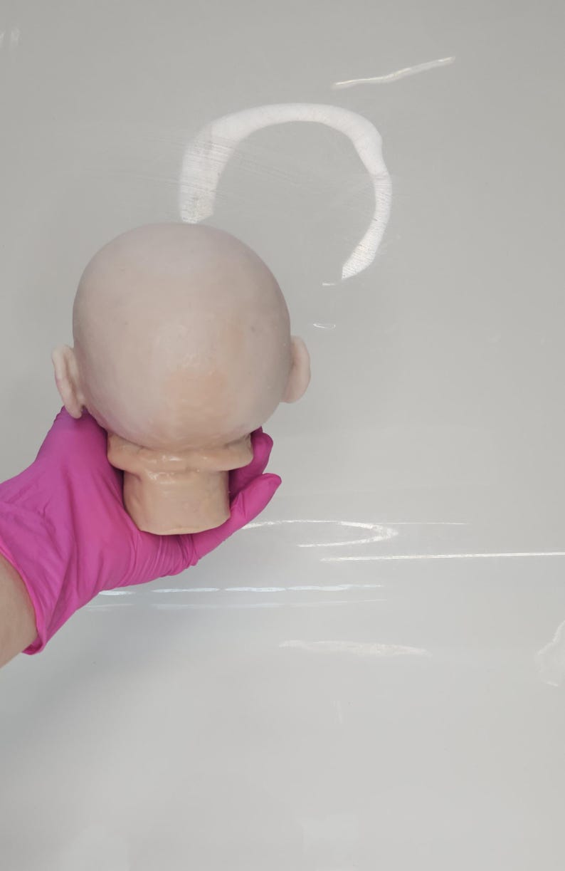 Pu&ograve; includere: Una testa di bambola calva e pallida con un cranio liscio e arrotondato e orecchie visibili. La testa &egrave; scolpita con un collo e una mascella definiti, caratterizzata da un tono di pelle chiaro con sottili variazioni di colore sul vinile.