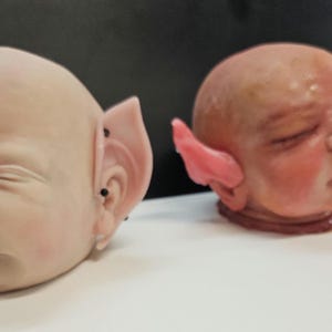 Può includere: Sono mostrate due teste di bambola, una con pelle chiara e piercing neri alle orecchie, l'altra con pelle più scura e un orecchio rosa. Le teste sono posizionate su una superficie bianca contro uno sfondo scuro. Le orecchie sono a punta.