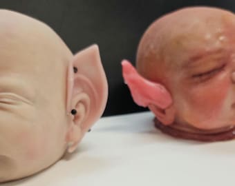 Puede incluir: Se muestran dos cabezas de muñeca, una con piel pálida y piercings negros en las orejas, la otra con piel más oscura y una oreja rosa. Las cabezas están colocadas sobre una superficie blanca contra un fondo oscuro. Las orejas son puntiagudas.