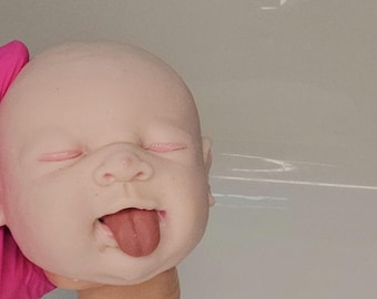 Puede incluir: Una cabeza de muñeco de piel pálida y lisa con ojos cerrados de párpados rosados, nariz pequeña y boca abierta con una lengua de color rosa oscuro que sobresale. La cabeza tiene forma redondeada y un cuello corto con rasgos realistas.