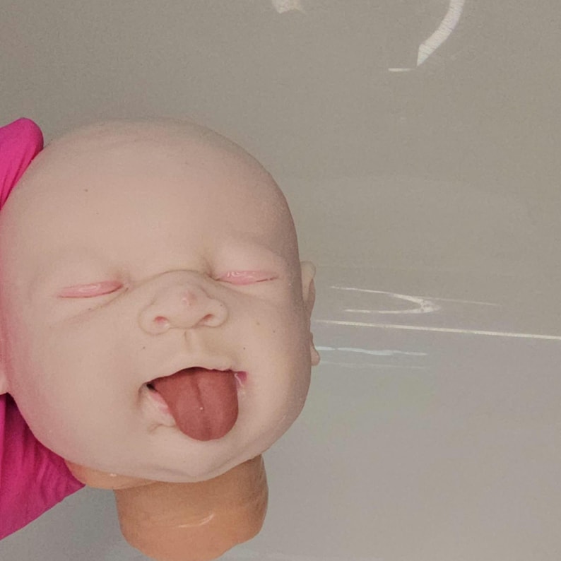 Pu&ograve; includere: Una testa di bambola dalla pelle pallida e liscia, con occhi chiusi dalle palpebre rosa, un naso piccolo e una bocca aperta con una lingua sporgente di colore rosa scuro. La testa ha una forma arrotondata e un collo corto dai tratti realistici.