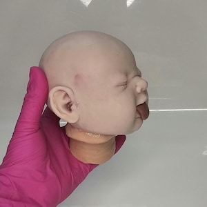Pu&ograve; includere: Una testa di bambola pallida e calva con gli occhi chiusi e una piccola lingua scura che sporge dalla bocca. La testa presenta tonalit&agrave; della pelle realistiche, un piccolo segno rosso sulla tempia e orecchie dettagliate, vista di profilo.