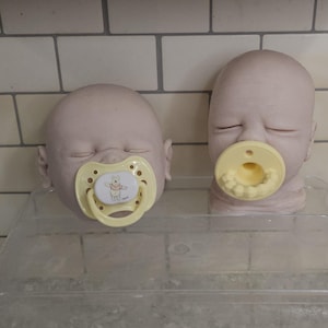 Pu&ograve; includere: Due teste di bambola pallide e calve con gli occhi chiusi. La testa a sinistra presenta un ciuccio giallo chiaro con un'immagine di Winnie the Pooh. La testa a destra presenta un ciuccio giallo chiaro con denti e gengive bianche modellate.